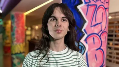 Kris Holland/BBC Harry Day with shoulder length dark hair. He has a nose ring and earring and is wearing a white T-shirt with thin green stripes on it.