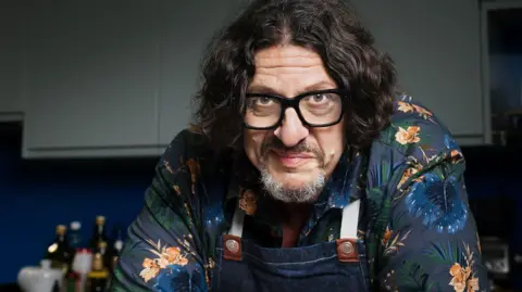 Jay Rayner Jay is looking leaning in towards the camera lens. He is wearing dark rimmed glasses and has mid-length dark brown hair. He is wearing a navy blue flowery short and denim apron and is stood in a kitchen.
