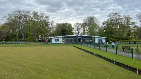 James Robinson/NCJ Media A bowling green with a single storey, blue building behind it. There is a path to the right of the green which has a couple of benches. There is another green to the right of the path, with a group of men gathered in the left corner. There are trees lining the back of the photo and the sky is grey with clouds.