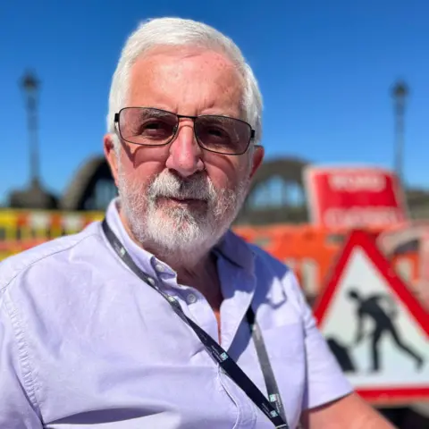 A man in a light purple shirt with grey hair and a grey beard and glasses is in front of road closure signs