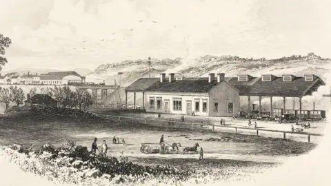 Getty Images A black and white engraving shows a single storey station building with four chimneys in the background. There is a steam train and carriages under a suspended roof above the platforms. In the foreground there are people milling about and three horses pulling a cart with a man riding at the front. 