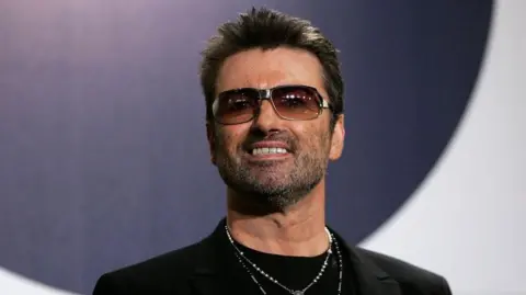 George Michael, who has short dark hair, a stubbly beard and dark sunglasses is looking ahead into the distance and smiling. He is wearing a black suit jacket and T-shirt and a necklace.  