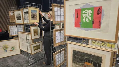 Leeds City Council A woman arranges framed artwork on a metal grid display. The display features multiple framed pieces, including a prominent abstract artwork with a central green panel flanked by red sections and a white organic shape in the middle, surrounded by black leaf-like motifs.