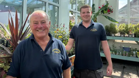 The two men with navy t-shirt standing in front of plants. Chris is older and stands in the front, with Alfie slightly behind him, both have short hair and are clean shaven. 