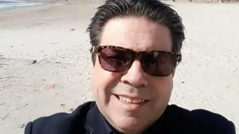 Grant Miller A middle aged man with black hair and tortoiseshell sunglasses smiles in a selfie, behind him is a beach with white sand. 