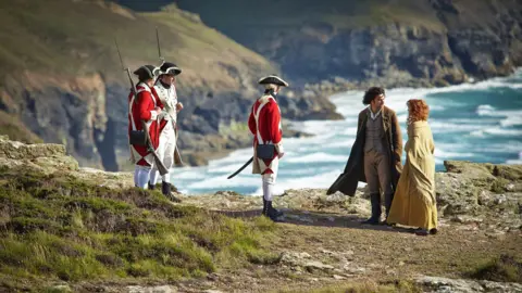 A woman with curly red hair and a beige trench coat stands next to a man with curly brown hair, a white shirt, brown waistcoat and trousers, and brown trench coat on the top of a cliff talking to each other. There are three soldiers in red and white uniform with tricorn hats holding bayonets standing near them. Beyond is a rugged coastline and rough sea.