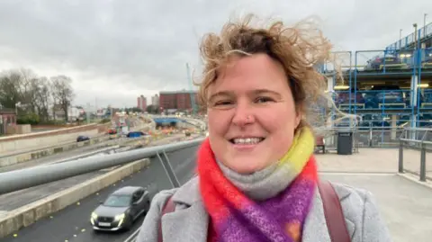 A woman smiling into the camera with curly windswept blonde hair, wearing a colourful scarf and grey blazer. You can see a road in the background with roadworks in the distance.