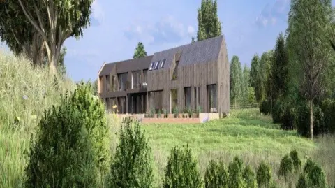 A tall brown house with large glass windows stands in the middle of a green field landscape that is surrounded by trees.