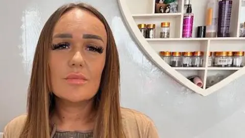 BBC Emma Bestall wears a sad expression as she looks into the camera. She has long brown hair and wears a cream cardigan. On the wall behind her, a heart-shaped shelving unit contains products from her hair and beauty salon.