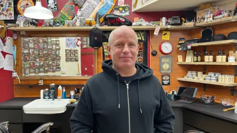 Holly Nichols/BBC The photo shows the interior of a barbershop filled with colourful decorations. Jason Keen is in the foreground wearing a black zip‑up hoodie. The barbershop wall is lined with shelves holding grooming products, jars, tins, and bottles. 