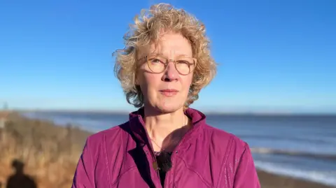 BBC Deborah Hawksley stands on the edge of the cliff at Skipsea. The sea is visible in the background with blue sky and grassed landscape. Ms Hawksley is wearing a dark pink jacket and gold necklace. She has light coloured glasses and blonde curly hair.