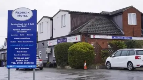 The front of the Redbeck Motel, as indicated by a blue and white sign in its car park. The sign plugs its food offering and features contact details. Two cars are parked outside the hotel, which is a relatively small two-storey building.
