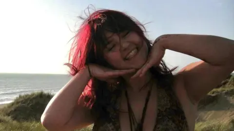 Mia Sanson-Regan Mia standing on a clifftop, smiling with the back of her hands under her chin.