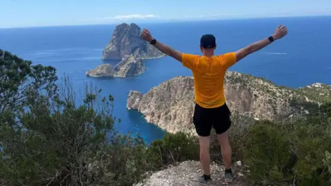 Mike Jones Mike Jones with his back to the camera, standing on an edge with his arms out looking at the sea and view in Ibiza in April 2025.
