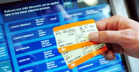 PA Media Two rail tickets being held in front of a ticket machine screen with various travel options and ticket types on display.