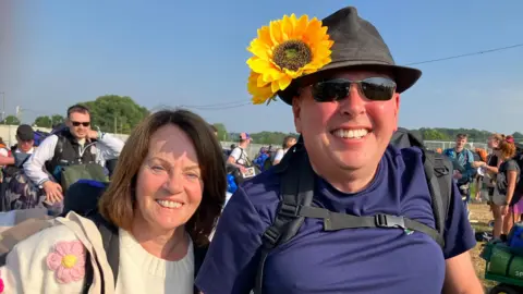 Carmel and Phil Cole pictured after entering Glastonbury Festival. Carmel wears a cream jumper with large crochet flowers, while Phil wears a dark hat with two large sunflowers attached to the brim. 