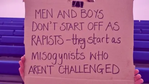 This Life Collective A cardboard sign is held in front of a girls face, her hands are visible on the edges. The phrase 'Rapists - they start as misogynists who aren't challenged' is written on the sign in black marker pen.