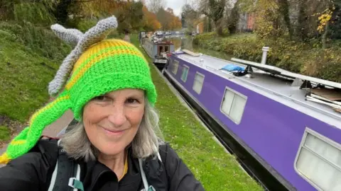 A woman with grey hair standing on a canal path wearing a knitted hat that resembles an air ambulance. A canal boat is next to her.