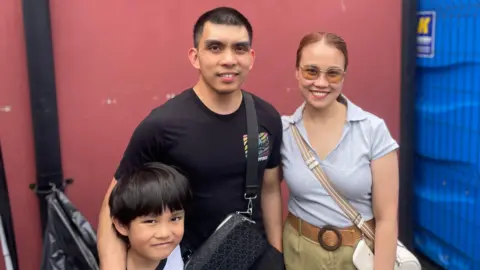 Three people stand in front of a red wall. There is a black metal fence beside them. A man with shirt black hair stands in the middle. He wears a black t-shirt and has a black bag over his shoulder. A small boy stands under the mans arm. He has short brown hair and a white t-shirt. To the mans left is a red-haired woman with a grey t-shirt, green trousers and white handbag. She wears orange lensed glasses. 