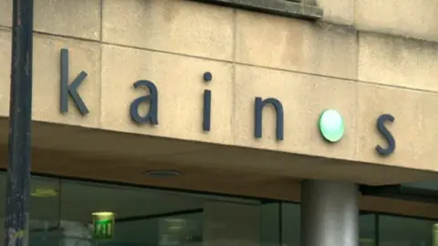 A shot of the exterior of Kainos's building in Belfast. The logo 'Kainos' written in lower case is in black with a green circle dot replacing the O.