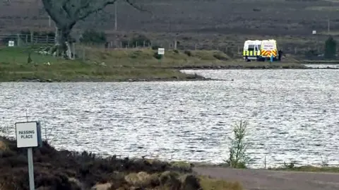 A calm loch with rippling water in the foreground, small evergreen trees along the near shore, and a lone person standing by the water’s edge across the lake. A white van with a yellow panel is parked on the far bank, with open moorland and low hills stretching into the background under an overcast sky.