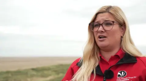 Amy Pennington, wearing a red Lancashire Wildlife Trust top with a badger logo, at the sand dunes in Lytham St Annes. She has long blonde hair and brown, round glasses.