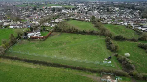People for Packsaddle An aerial view of a field with a metal fence partitioning off a muddy path along a hedgerow. There are houses around more fields in the background.