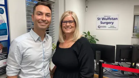 A man and woman stand in a doctors' surgery. He has brown hair and is in a pale blue shirt and she is blonde and wearing glasses and a black dress.  A sign on the wall behind them says Welcome to St Wulfstan Surgery, Care Quality Commission Rated Outstanding and computers can be seen on desks.