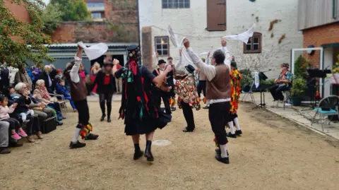 The Folk of Gloucester A group of Morris dancers in traditional costume are waving handkerchiefs in the air. An accordion player is in the middle of the circle of dancers wearing a top hat and black 'tatter' coat covered in ribbons.