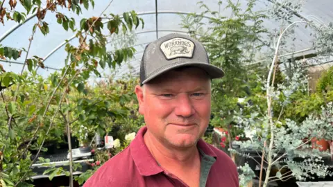 John Devine/BBC John Cutteridge, 51, has a baseball cap on with no hair visible and a mauve t-shirt. Behind him are lots of plants and shrubs, inside a large poly tunnel