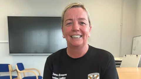 Avoureen Coleman has blonde hair in a ponytail. She's wearing stud earrings and a black top that says Clann Éireann senior ladies on the right hand side and the Clann Éireann white and gold logo on the left hand side. Behind her is a white wall with a large TV screen.