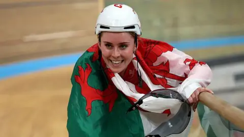 Emma Finucane, with a Wales flag around her neck, celebrates a gold medal at the Commonwealth Games
