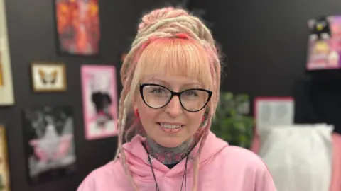 A lady with multiple piercings and black glasses. She has blonde hair and dreadlocks and shades of pink in her hair. 