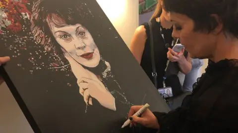 Sue Verity Art A painting of Helen McCrory is being signed by Helen McCrory, a woman in a black top and short hair is signing a portrait of herself