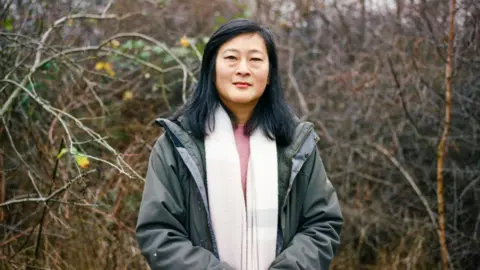 Joanne Coates Head and shoulder view of Emi Imai, who has shoulder-length black hair in a side parting. She is wearing a grey cagoule and a long white scarf over a pink jumper. Behind her is mostly bare undergrowth, with a few yellow flowers - possibly daffodils.