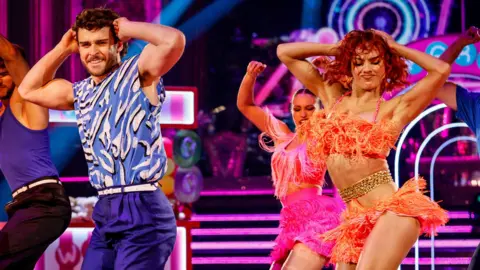 PA Media George Clarke wears a blue and white top with purple trousers and dances next to his partner Alexis Warr who wears an orange two piece with frills, they have their hands on their head as they dance and there are three backing dancers behind them.