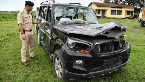 AFP via Getty Images This photo taken in July 2018 shows Gulshan Daolagupu, deputy superintendent of police (in uniform), showing the damaged vehicle in which two men were lynched in Assam's Karbi Anglong district. The car is a black Scorpio SUV, with its bonnet crashed, windshield broken, doors dented.
