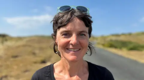 Pip Cook has short brown hair. She has blue sunglasses on her head and gold hoop earrings. She is wearing a black top. In the background is a road and grass. The sky is blue.
