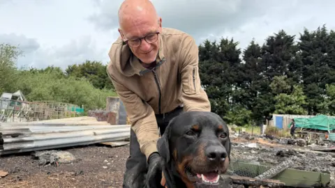 Martin Taylor with his hands around his Rottweiler dog who is standing in front of him. Martin has a shaved head and is wearing glasses and a beige coat with dark trousers