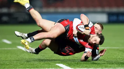 Getty Images Two rugby players tackling each other to the ground with a rugby ball in hand.