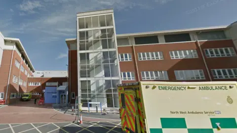 Nondescript brown hospital building with large windows with green ambulance in front.