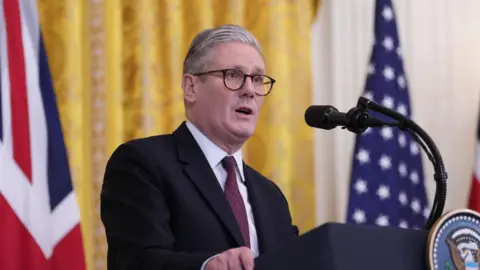 Sir Keir Starmer in a suit and tie stands at a podium in the White House with the presidential seal, flanked by British and American flags.