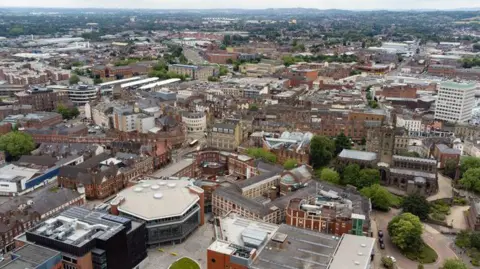 An aerial view over the city of Wolverhampton on an overcast day. There are old factory buildings and new developments, with hills in the background.