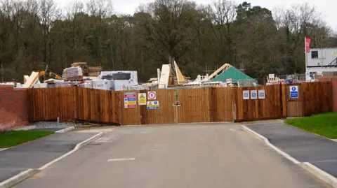 Qays Najm/BBC Tall wooden slat fencing, with construction notices, runs across a road. There are pallets and building materials behind the fencing.