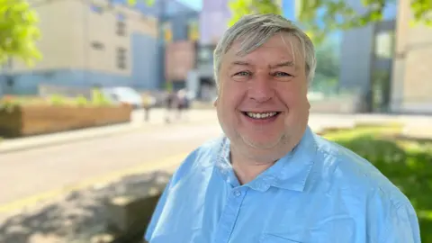 Jon Hubbard smiles at the camera. He wears a blue shirt - in the background, the blurred Wiltshire council HQ on a sunny day