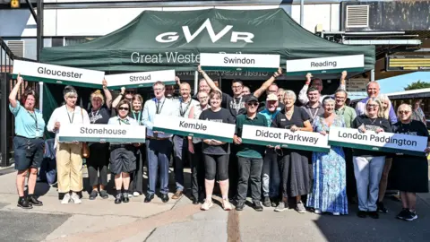 Great Western Railway A large group of Great Western Railway staff smile and celebrate as they stand on a railway platform, holding signs bearing the names of various stations on the Gloucester to London Paddington route, including Kemble and Didcot Parkway. It is a sunny day and they are stood in front of a dark green Great Western Railway marquee.