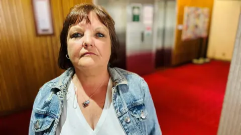 Woman - Lynn Winstanley - looking at camera inside a council building, she is wearing a white v-neck top under a light blue denim jacket, and a necklace.
