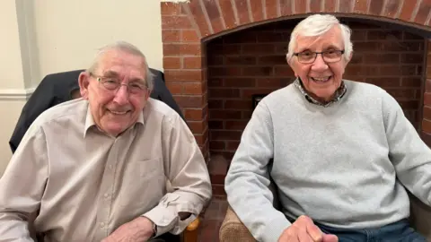 BBC The photo shows two elderly men smiling at the camera. They both have glasses and grey hair. The one on the left is wearing a brown checked shirt. The one on the right has a grey jumper with a shirt underneath. They are sitting in armchairs, in front of a fireplace. 