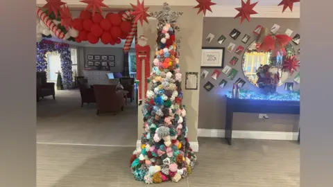 Grampian Court Care Home The Christmas tree is made of fluffy pompoms in different sizes and colours. A big silver star is placed at its top. A Santa Claus figure is handing on the wall next to it,.The rest of the room is decorated with lights. Red stars are handing from the ceiling and Christmas cards are attached to one of the walls. 