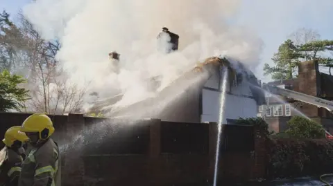 Thatched cottage with its roof on fire and tick smoke billowing out. Two firefighters are seen to the left and several hoses are spraying water onto the roof.